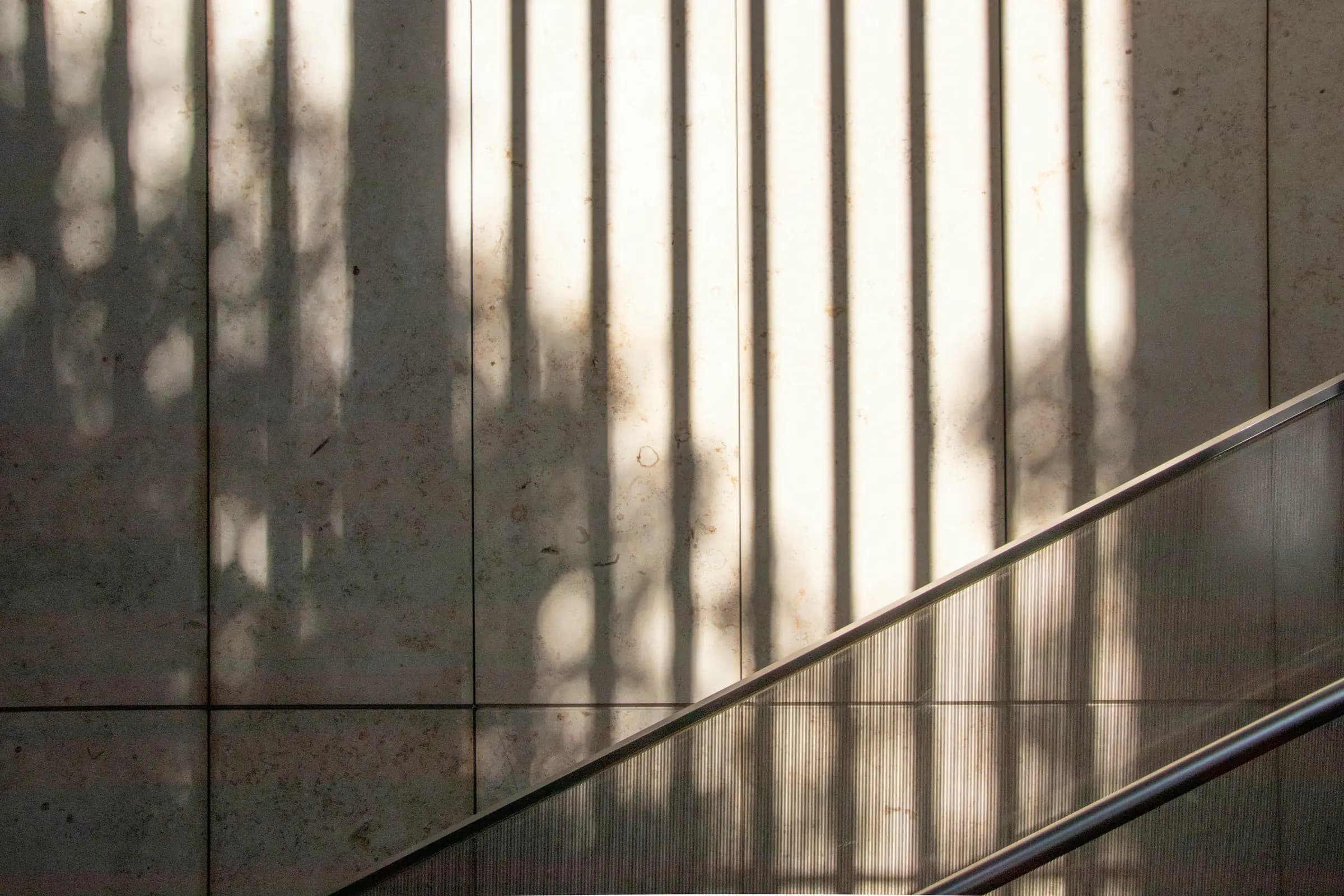 Vertical bands of light and shadow falling across a pale stone wall beside a stair handrail